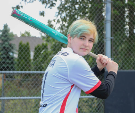 Emma Kerani, blue hair and all, holding a softball bat over their shoulder out on the softball field and getting ready to swing.