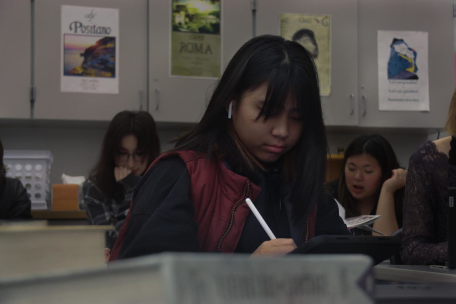A posed photo of Arabella Devera looking down at her work with a solemn face, holding a pen and listening to music. In the background, other students are blurred, also working on their own schoolwork.