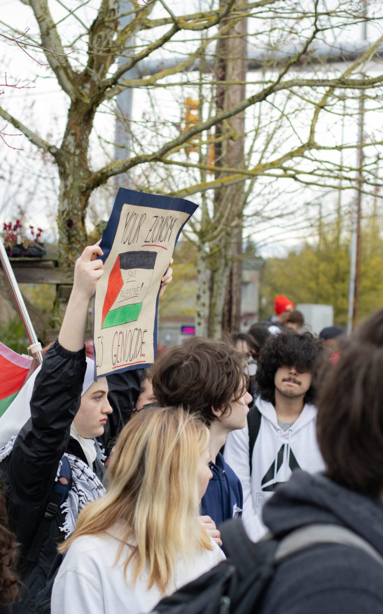 Students gather along the sidewalk, with one holding up a sign saying Your zionism is genocide.